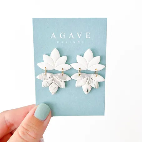 A hand with mint green nail polish holds a blue card featuring the brand name "Agave Designs." Attached to the card is a pair of sterling silver Jodie earrings, designed with layered leaves and small marble-patterned leaves hanging below.