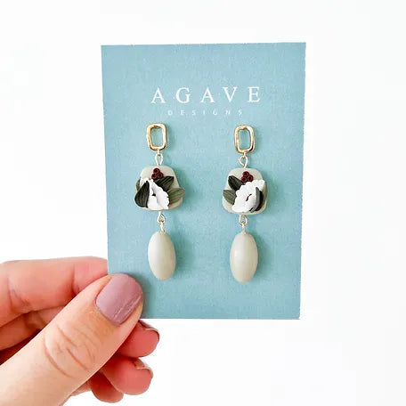 A hand with mauve-painted nails holds a light blue card displaying a pair of Noelle drop earrings. The earrings from Agave Designs feature a gold-plated square stud, a floral design in white and burgundy, and an attached oval-shaped pendant in muted cream. The card reads "Agave Designs.