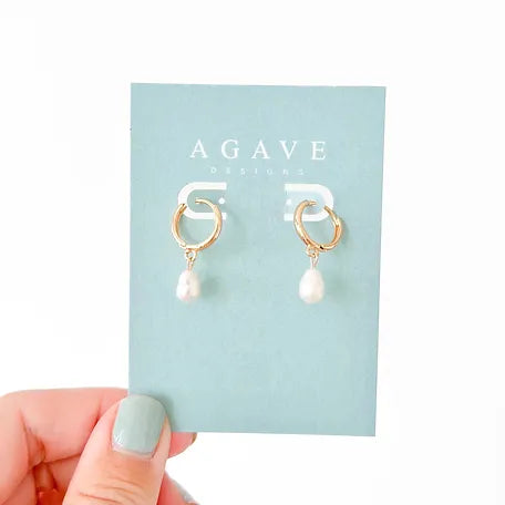 A hand with light blue nail polish holds a card displaying a pair of Pearl Huggies by Agave Designs, featuring gold hoops with dangling natural freshwater pearls. The card has "AGAVE DESIGNS" printed on it in white text.